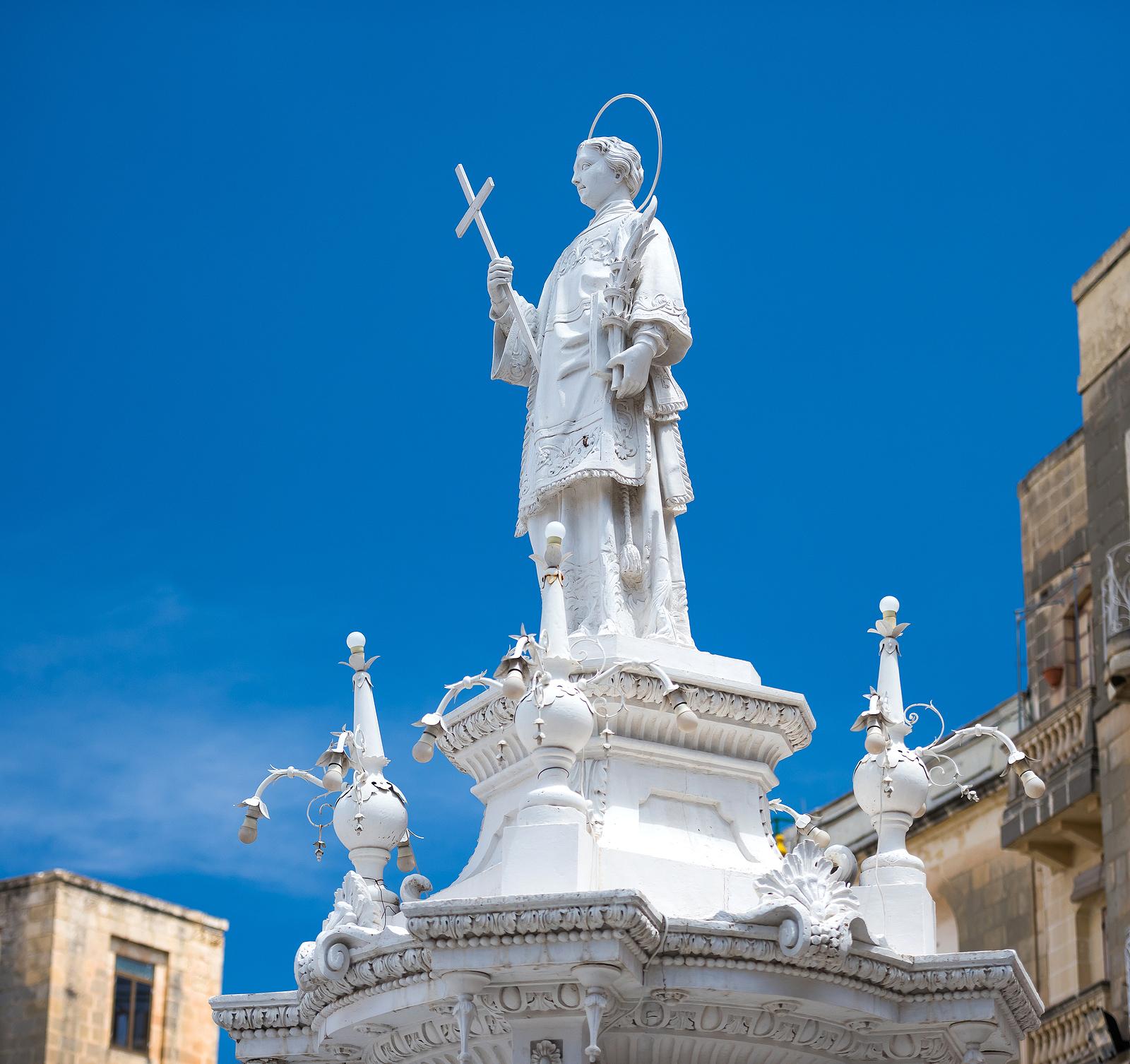 Statue of St. Lawrence in front of a blue sky. He is holding a cross.